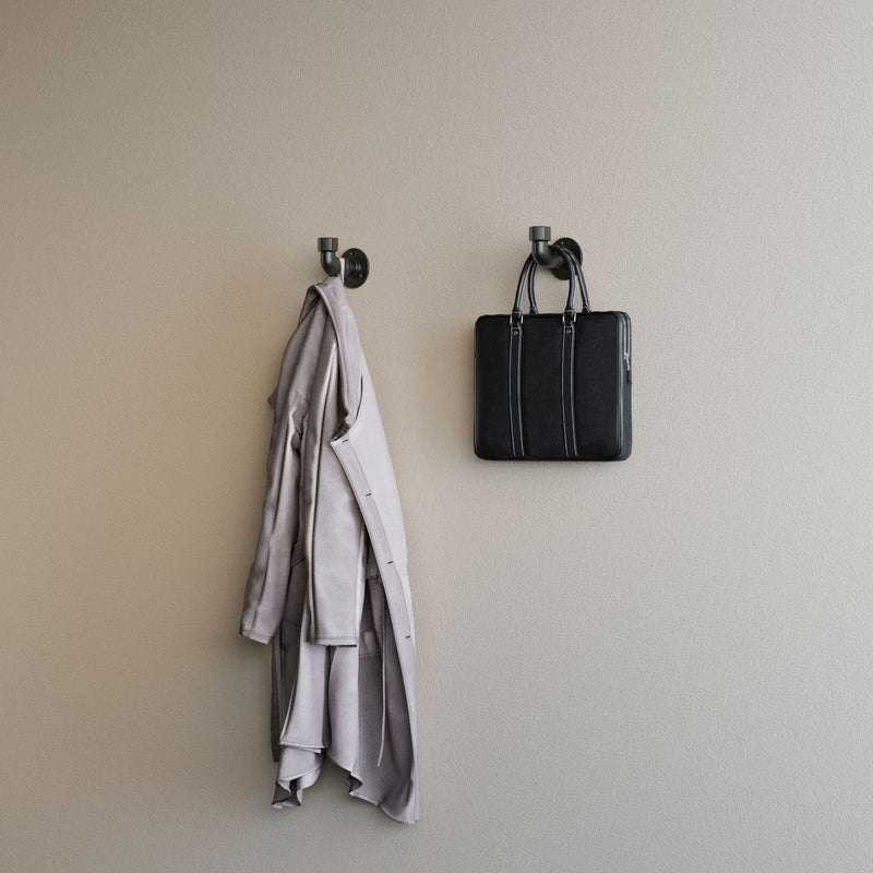 Hellbeige Wand mit zwei schwarzen Kleiderhaken, an einem hängt eine graue Jacke, am anderen eine schwarze Tasche